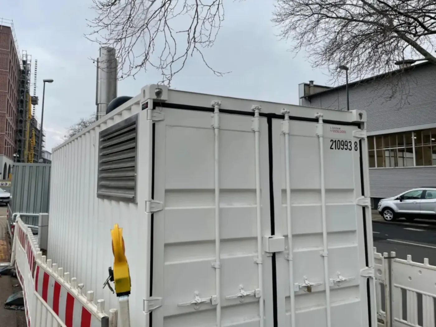 Weisser Heizcontainer mit Abgaskamin am Rand einer Baustelle in der Stadt, abgesperrt mit rot-weissem Bauzaun