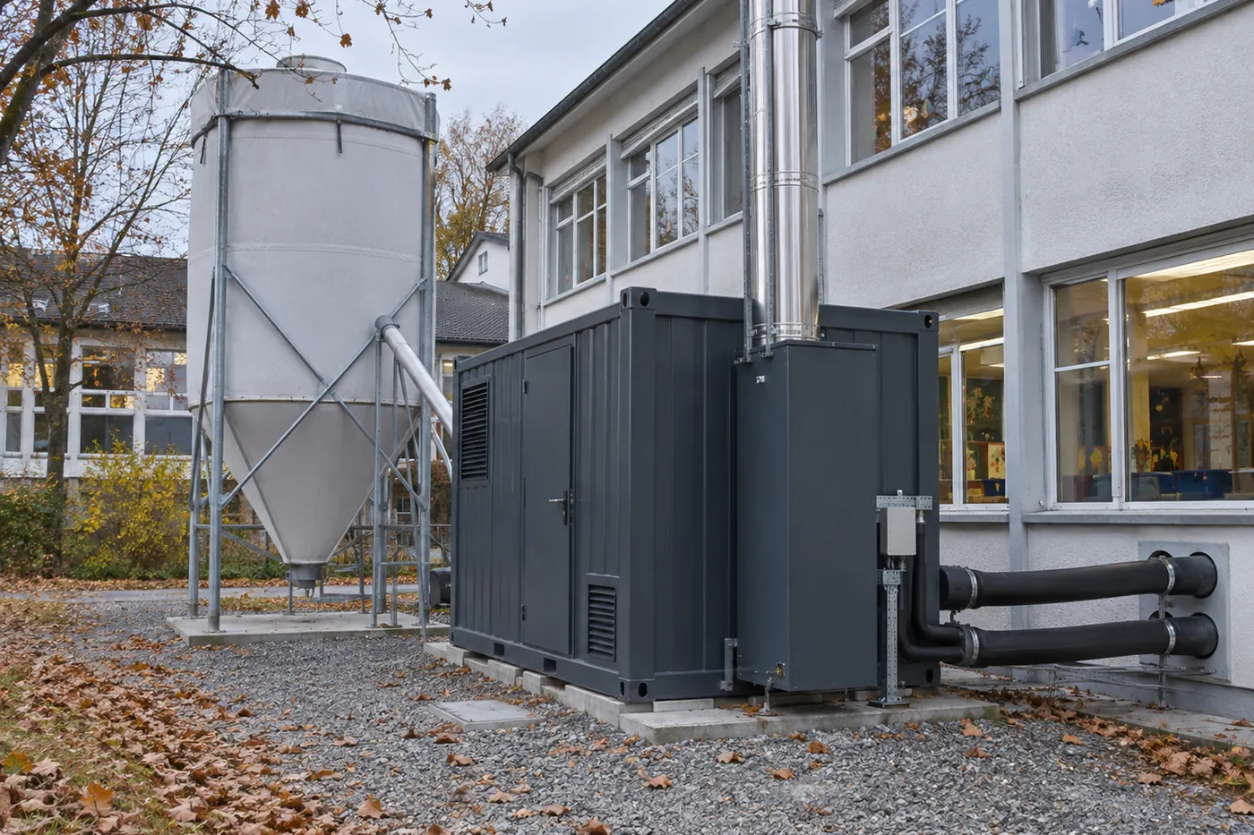 Anthrazitgrauer Heizcontainer mit Edelstahlkamin neben einem Pelletsilo auf Stahlgestell vor einem Schulhaus, isolierte Leitungen führen ins Gebäude