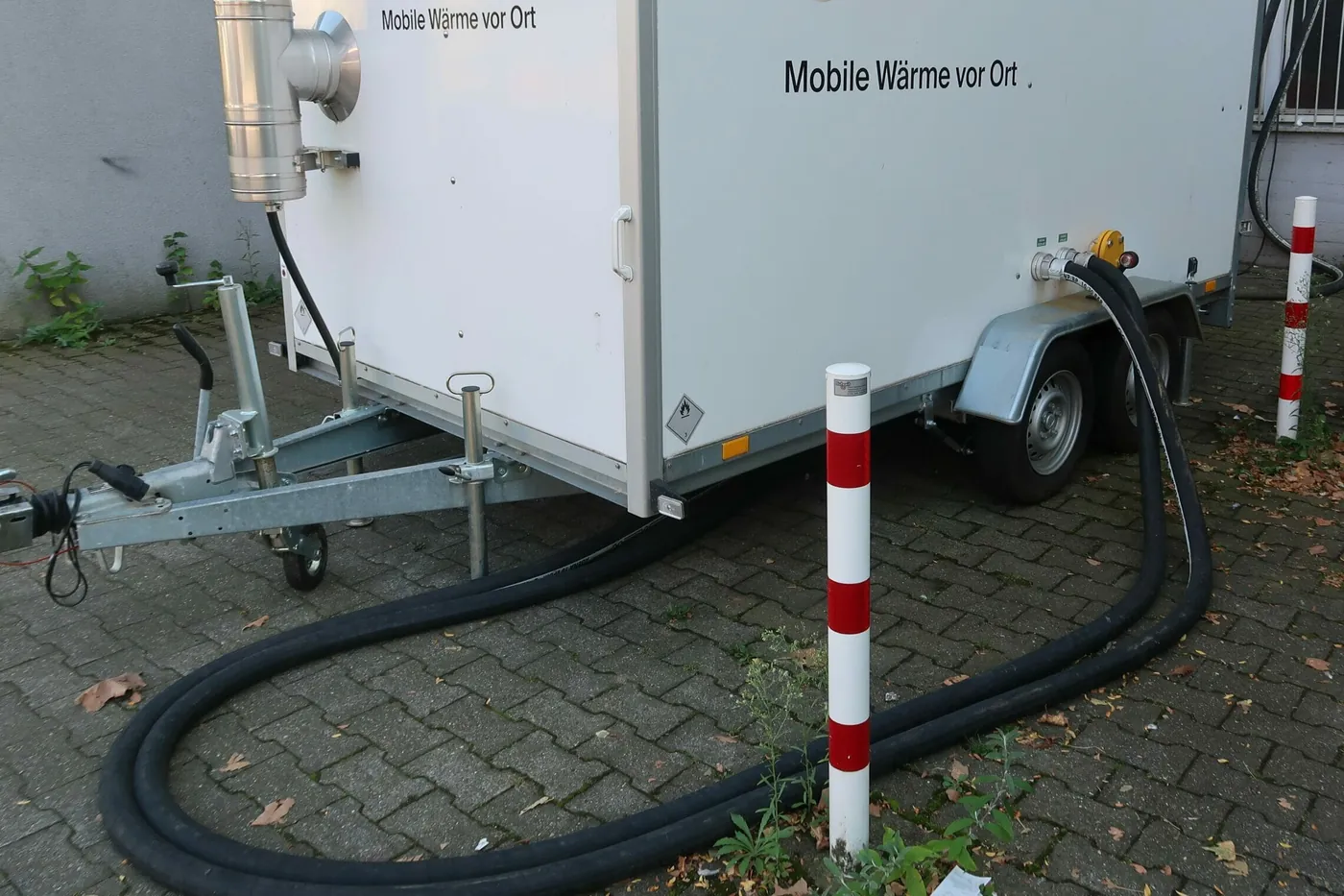 Heizmobil im weissen Kofferanhänger mit Edelstahlkamin, zwei schwarze Heizschläuche führen über das Pflaster zum Gebäude