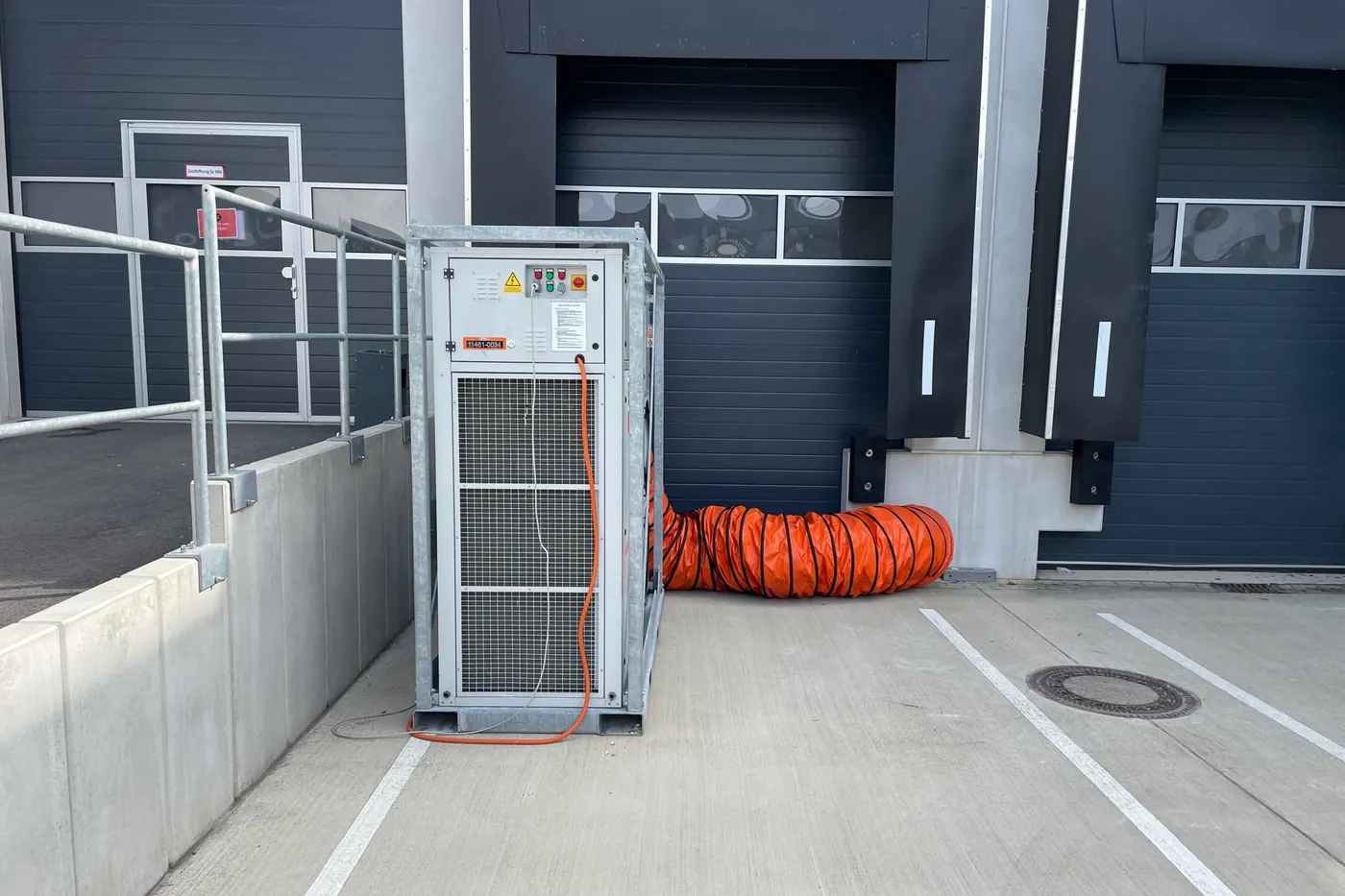 Mobile Klimaanlage im Stahlgestell an der Laderampe einer Halle, daneben ein orangefarbener Luftschlauch vor dem Hallentor
